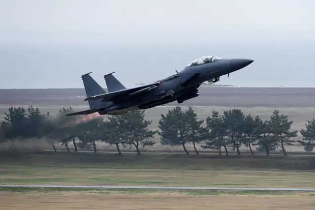 An F-15K lifting off with afterburners lit, coastline visible beyond the airfield