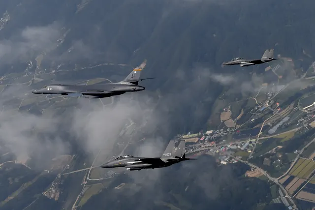 A B-1B bomber flying with two F-15Ks above farmland and wooded hills