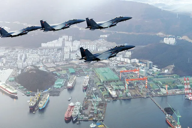 Four F-15Ks flying above a shipyard with ships alongside and gantry cranes