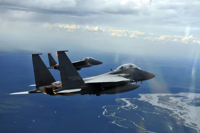 Two F-15Ks in flight above forest and a braided river, seen from another aircraft